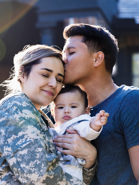 Female servicemember in uniform holding her young child while husband kisses her on the head.